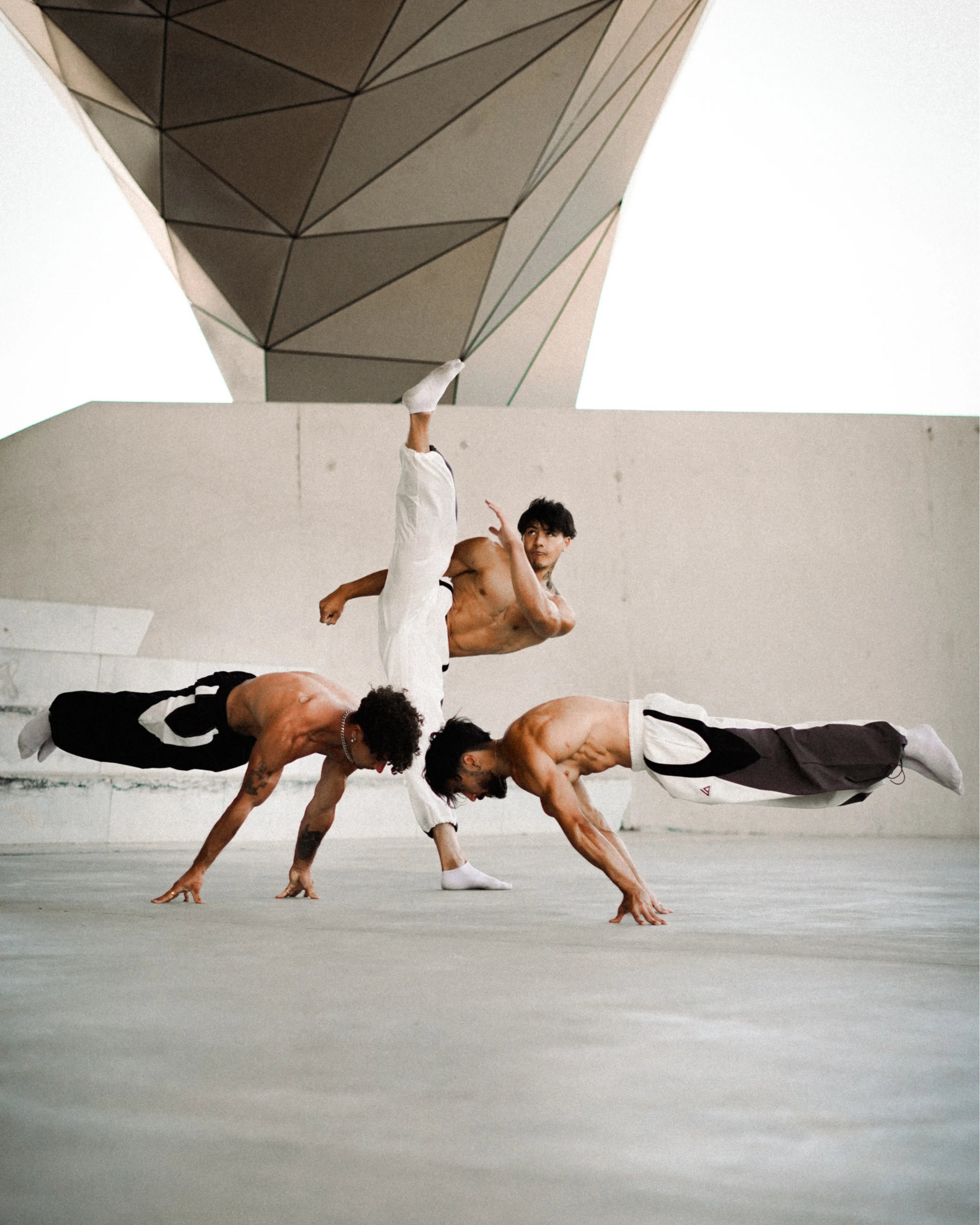 Three shirtless martial artists in white and black uniforms perform dynamic acrobatic karate poses in a large concrete courtyard under a geometric roof structure.