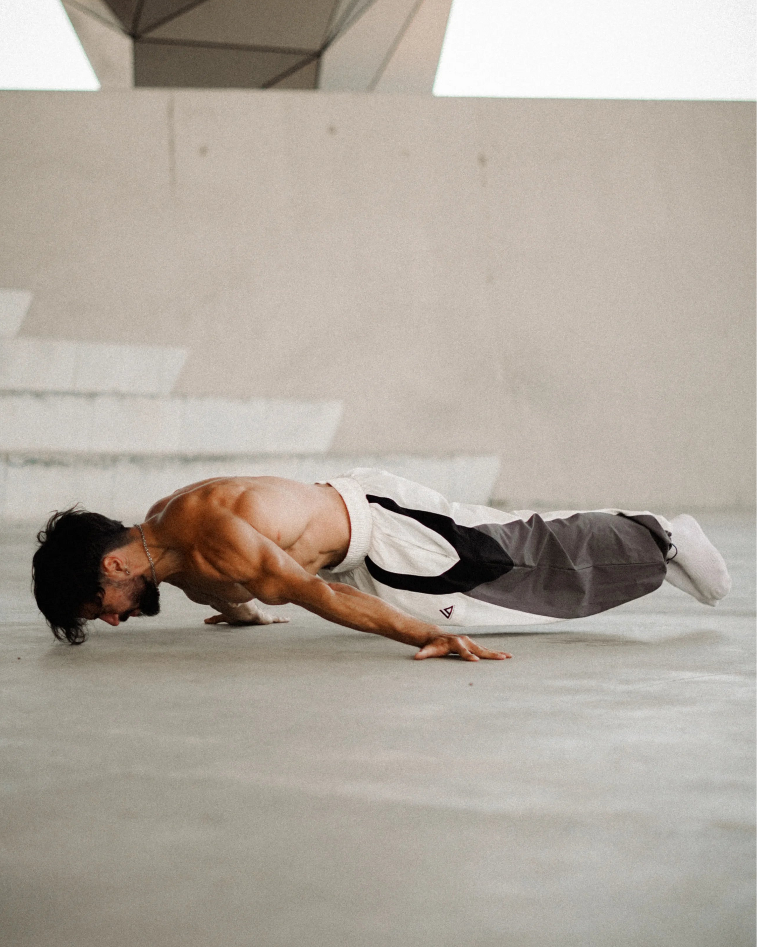 A shirtless man in a martial arts uniform performing a push-up on a concrete floor in front of a geometric modern building.