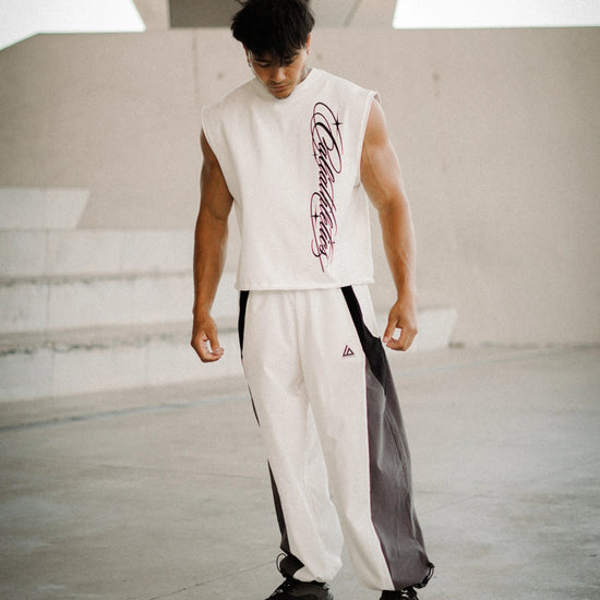 A man wearing a sleeveless white athletic top with a purple graphic print and matching white track pants with black side panels, standing in a modern concrete architectural setting.