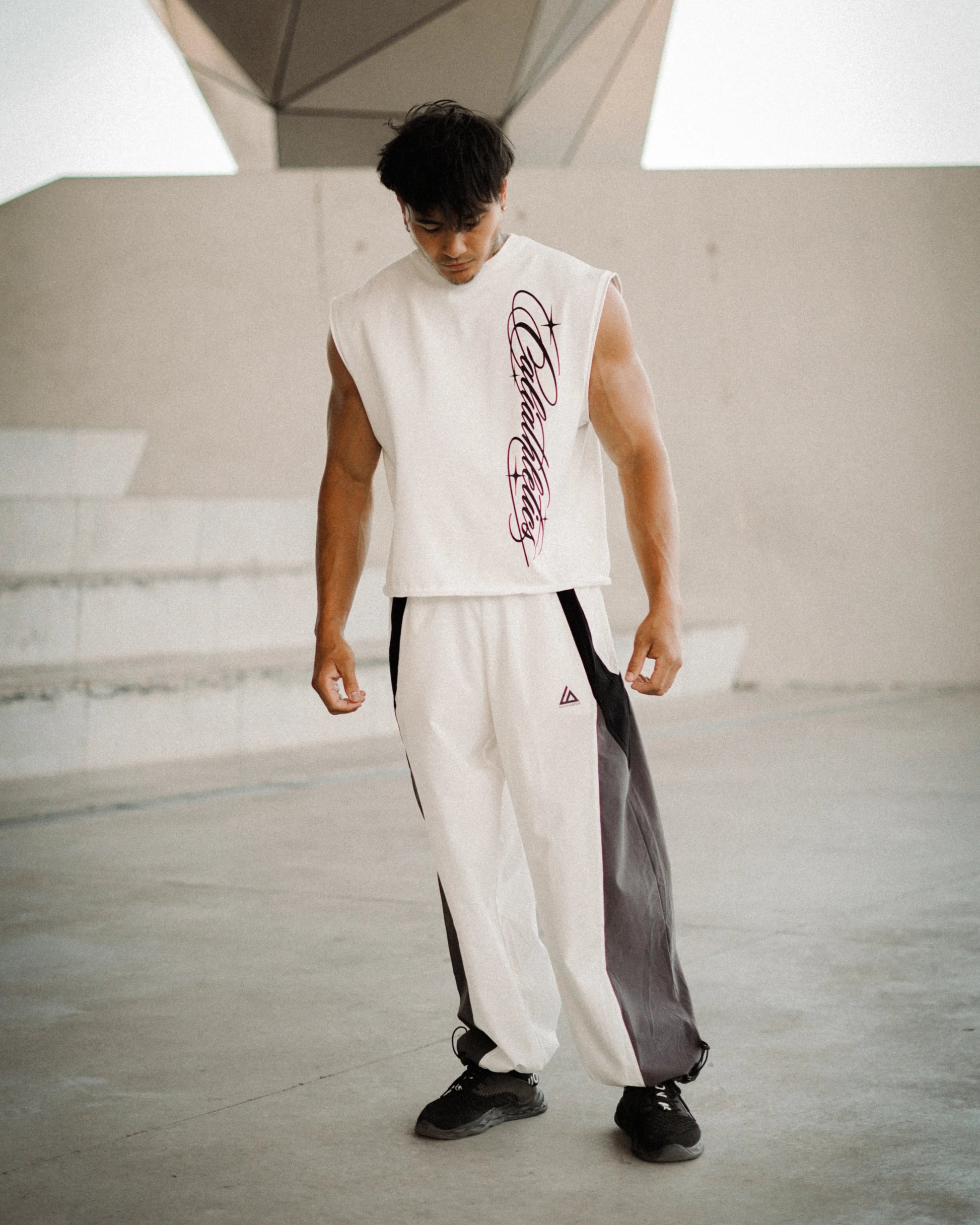 A man wearing a sleeveless white athletic top with a purple graphic print and matching white track pants with black side panels, standing in a modern concrete architectural setting.