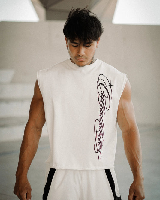 A muscular man wearing a sleeveless white athletic top with a black graphic print on the front, paired with white track pants with black side stripes, standing in a modern concrete structure.
