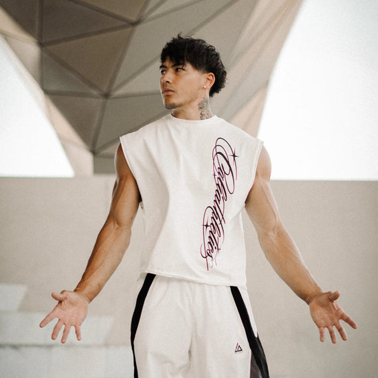 A man wearing a sleeveless white athletic top with a purple graphic print and matching white track pants with black side panels, standing in a modern geometric concrete structure.