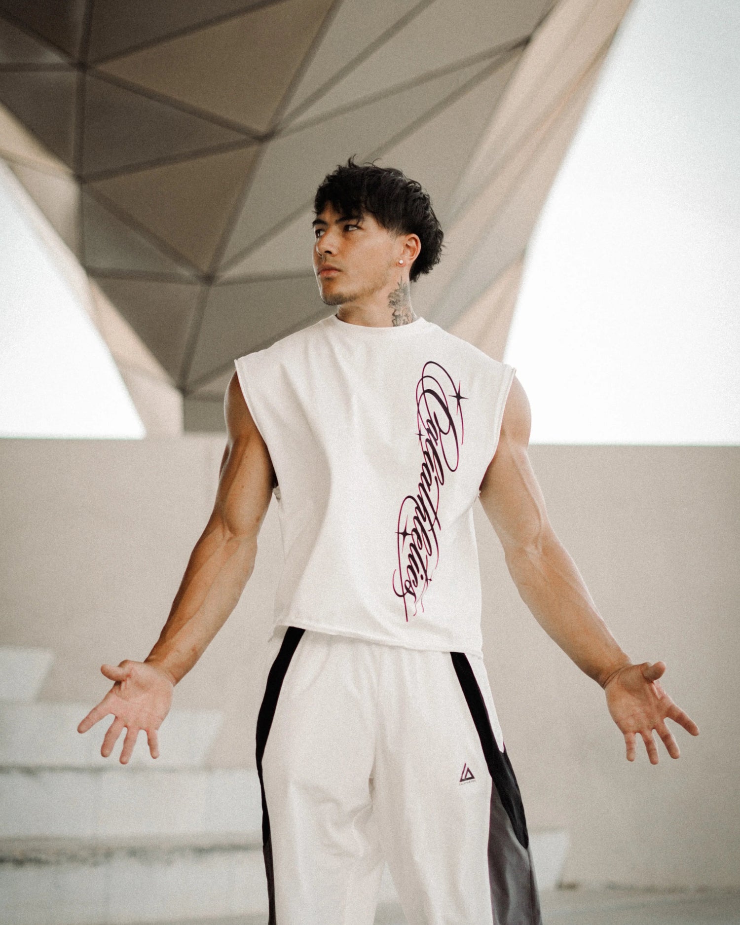 A man wearing a sleeveless white athletic top with a purple graphic print and matching white track pants with black side panels, standing in a modern geometric concrete structure.