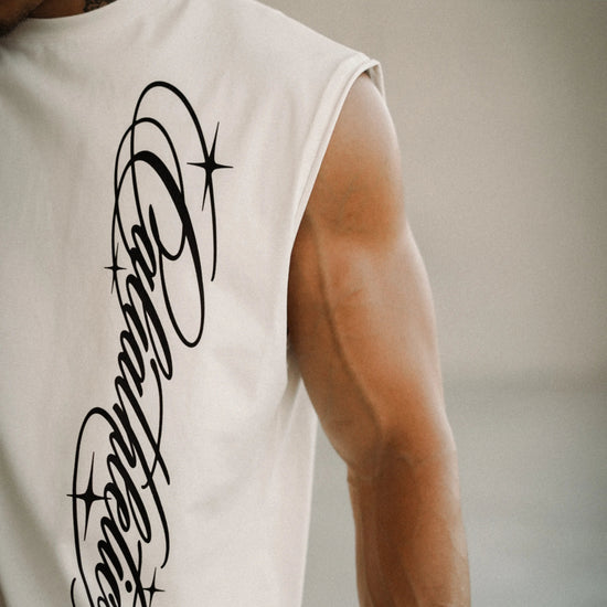 A close-up of a man's muscular arm and shoulder wearing a sleeveless white athletic tank top with a large black script graphic on the back, set against a blurred indoor background.