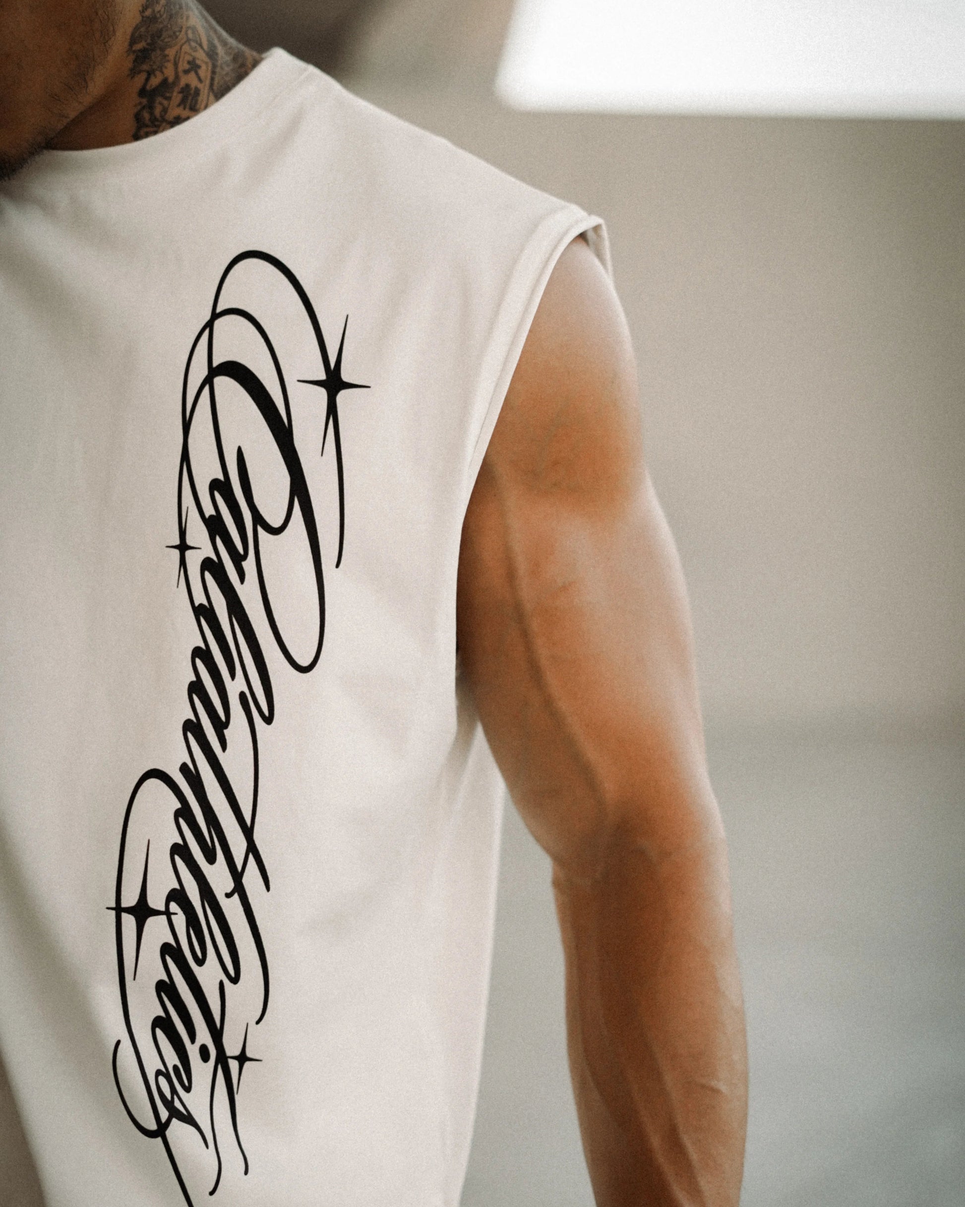 A close-up of a man's muscular arm and shoulder wearing a sleeveless white athletic tank top with a large black script graphic on the back, set against a blurred indoor background.