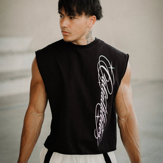 A muscular man wearing a black sleeveless crop top with a white graphic print on the side, paired with white athletic shorts, standing in a modern concrete structure.