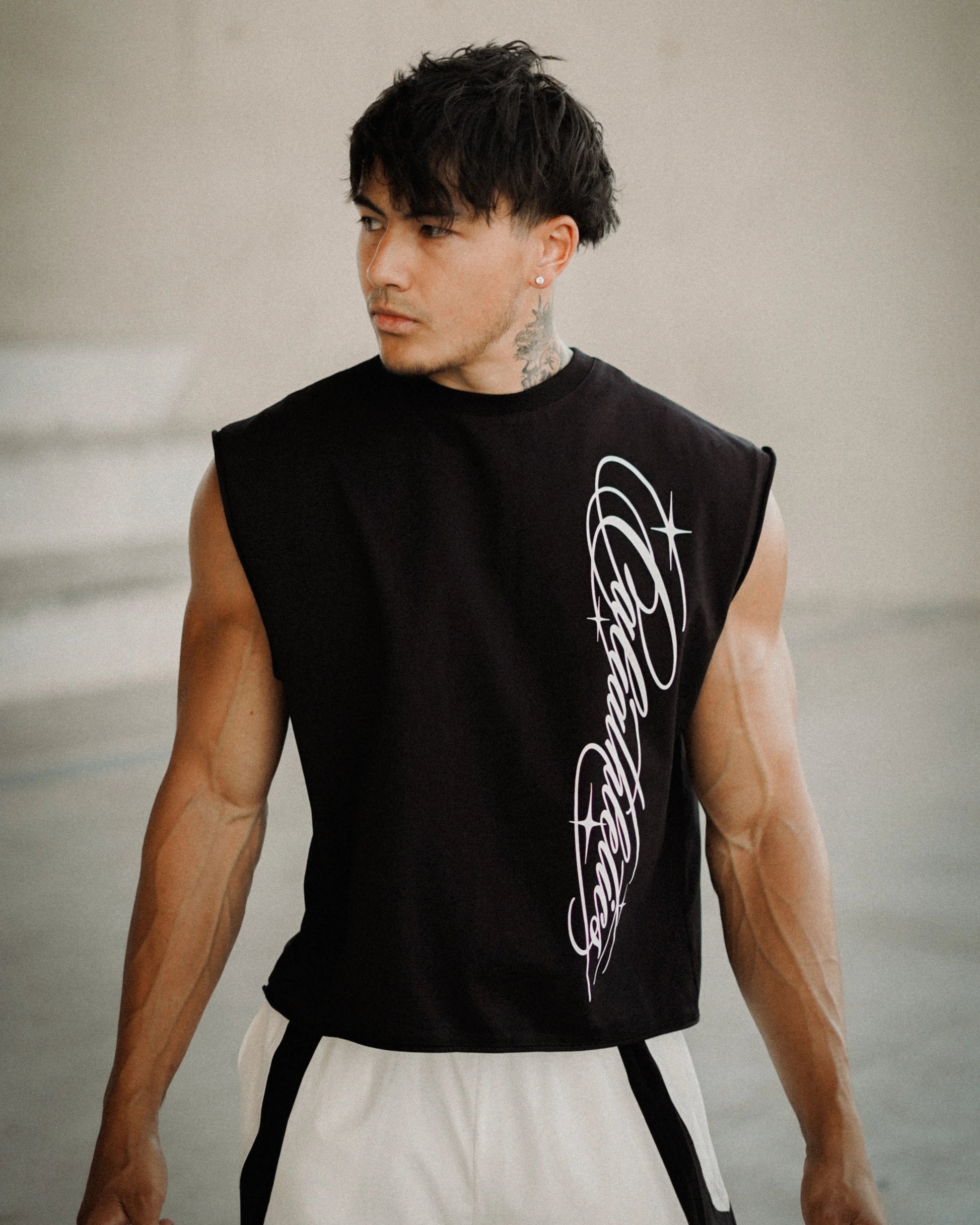 A muscular man wearing a black sleeveless crop top with a white graphic print on the side, paired with white athletic shorts, standing in a modern concrete structure.
