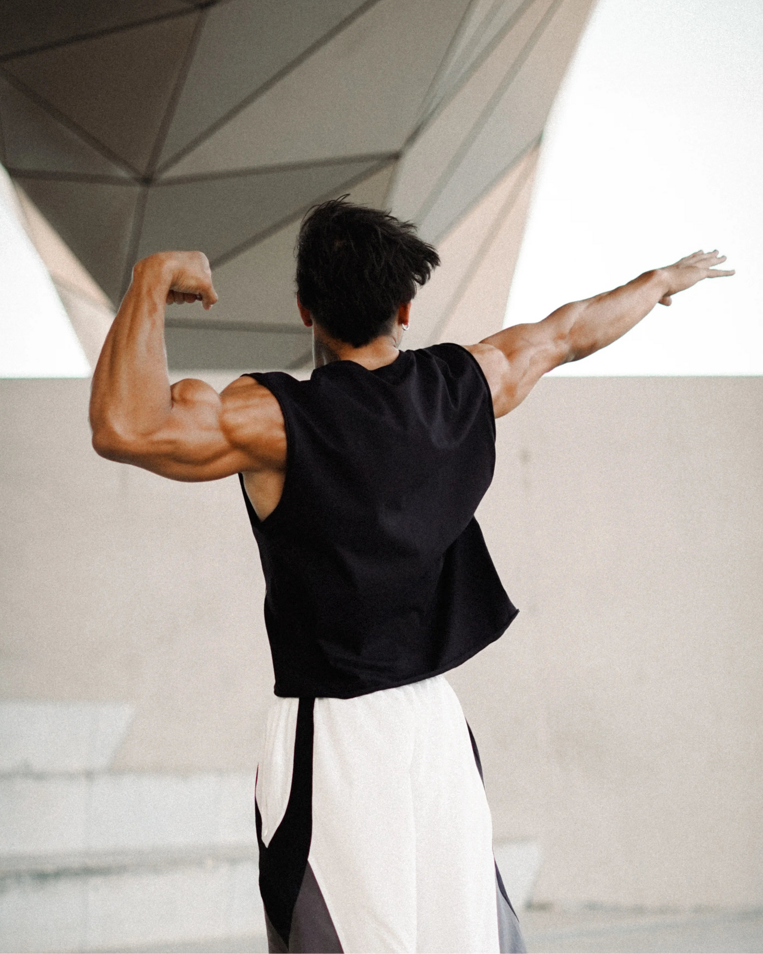 A muscular man shown from behind, flexing his bicep while wearing a black sleeveless shirt and white, gray, and black athletic pants in a modern concrete structure.