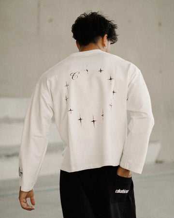 A man viewed from behind wearing a white long-sleeve crop top with black graphic prints on the back and black wide-leg sweatpants, standing in a modern concrete architectural setting.