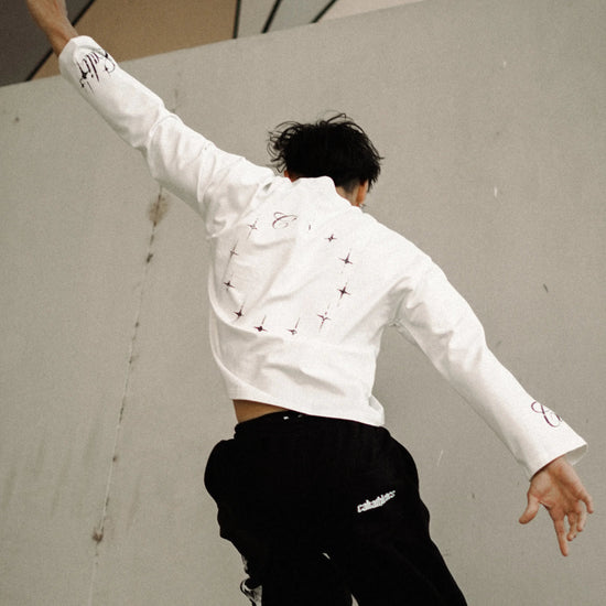 A person wearing a white long-sleeve cropped shirt with small black details and black wide-leg pants is captured mid-motion against a large concrete wall in an outdoor urban setting.