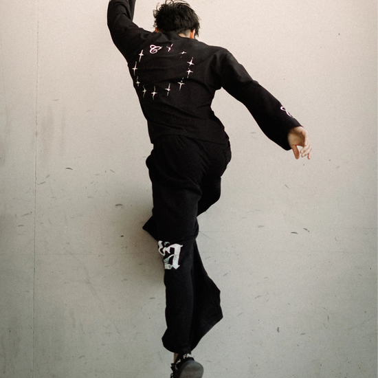 A person in a black outfit with white graphic details is captured mid-jump against a plain light gray wall.