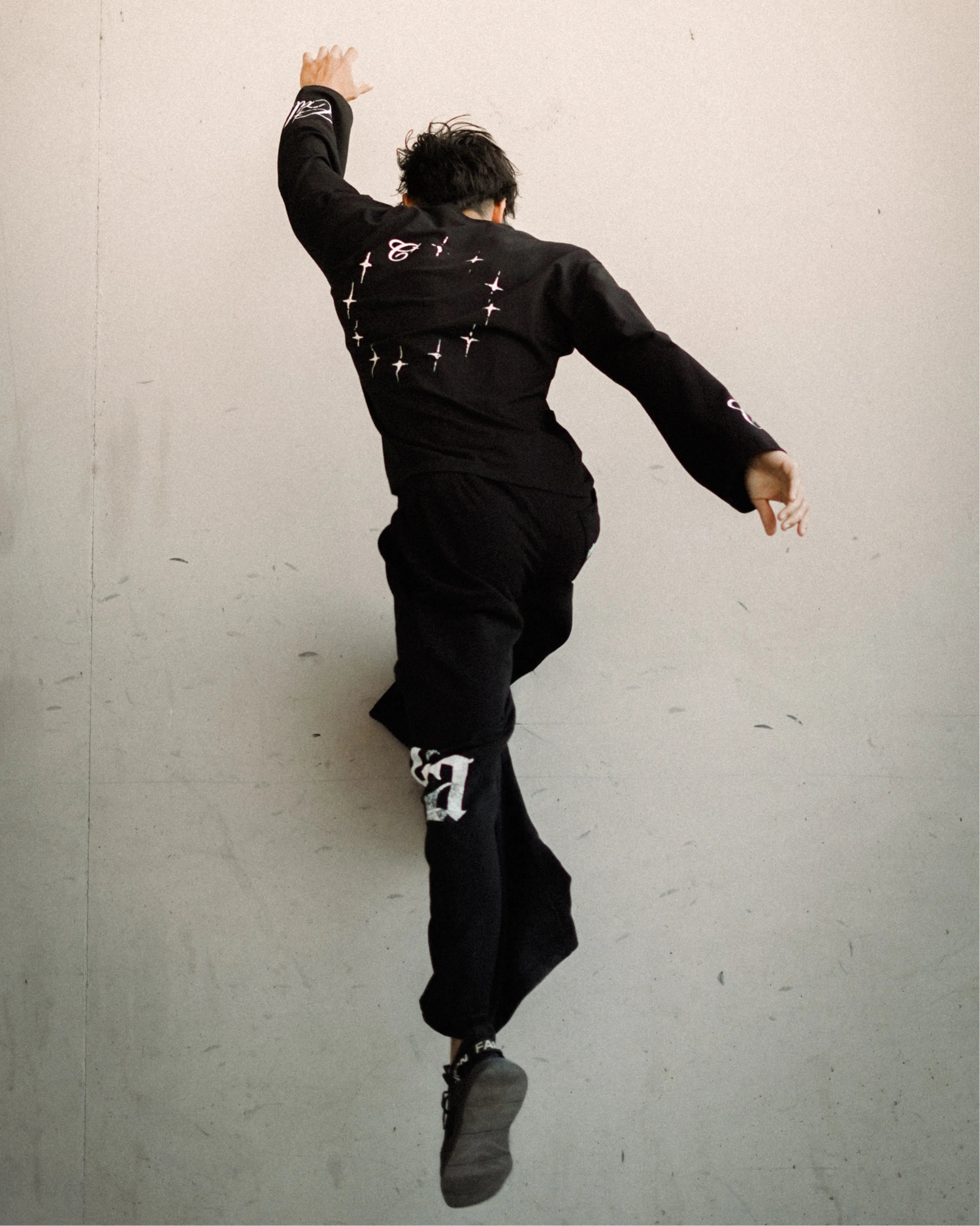 A person in a black outfit with white graphic details is captured mid-jump against a plain light gray wall.